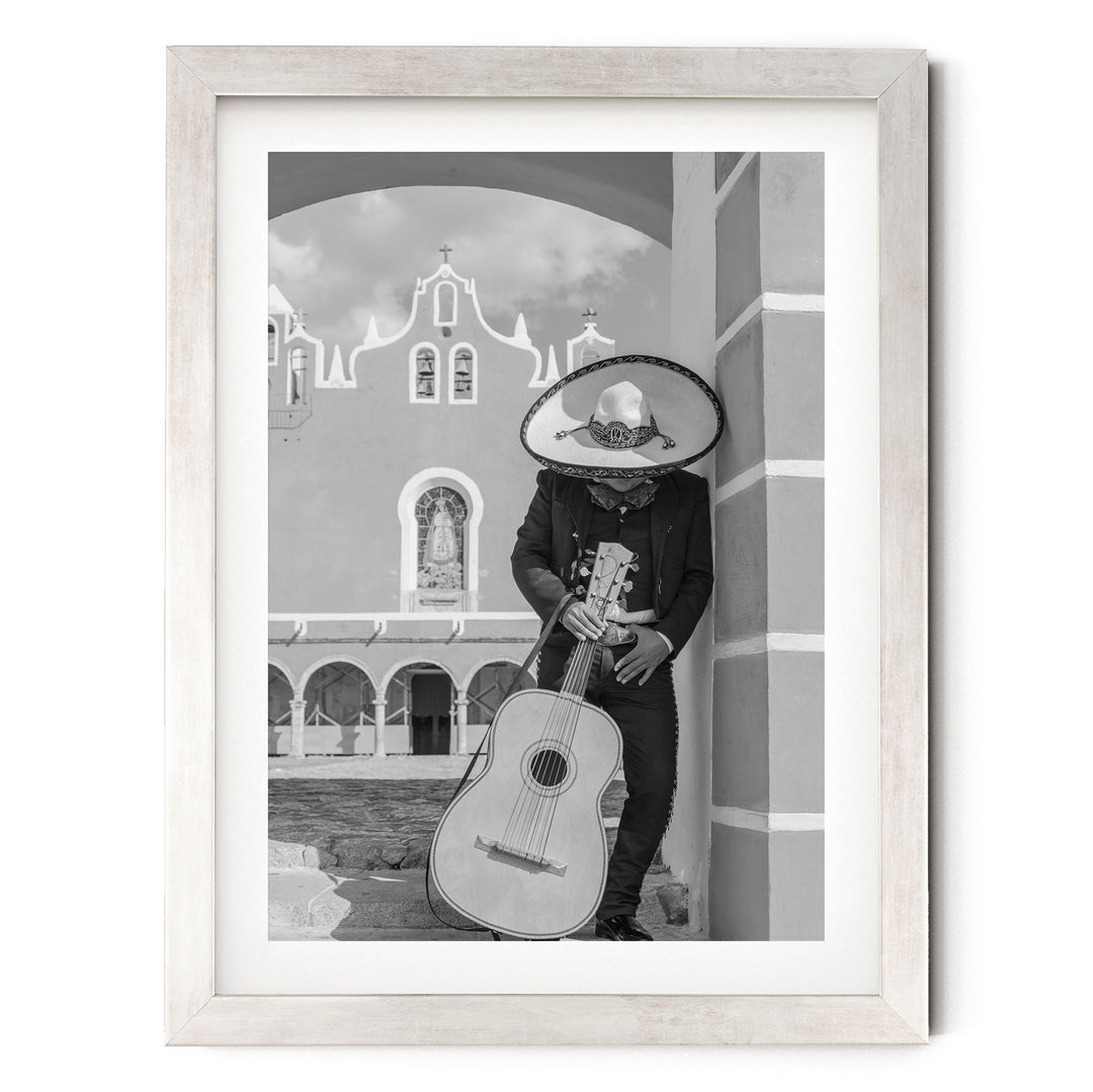 A person in a sombrero and traditional Mexican attire is playing a guitar in front of a building with arched windows and a bell tower.