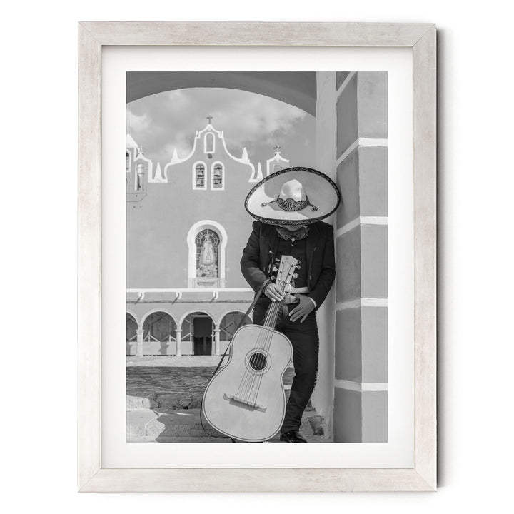 A person in a sombrero and traditional Mexican attire is playing a guitar in front of a building with arched windows and a bell tower.