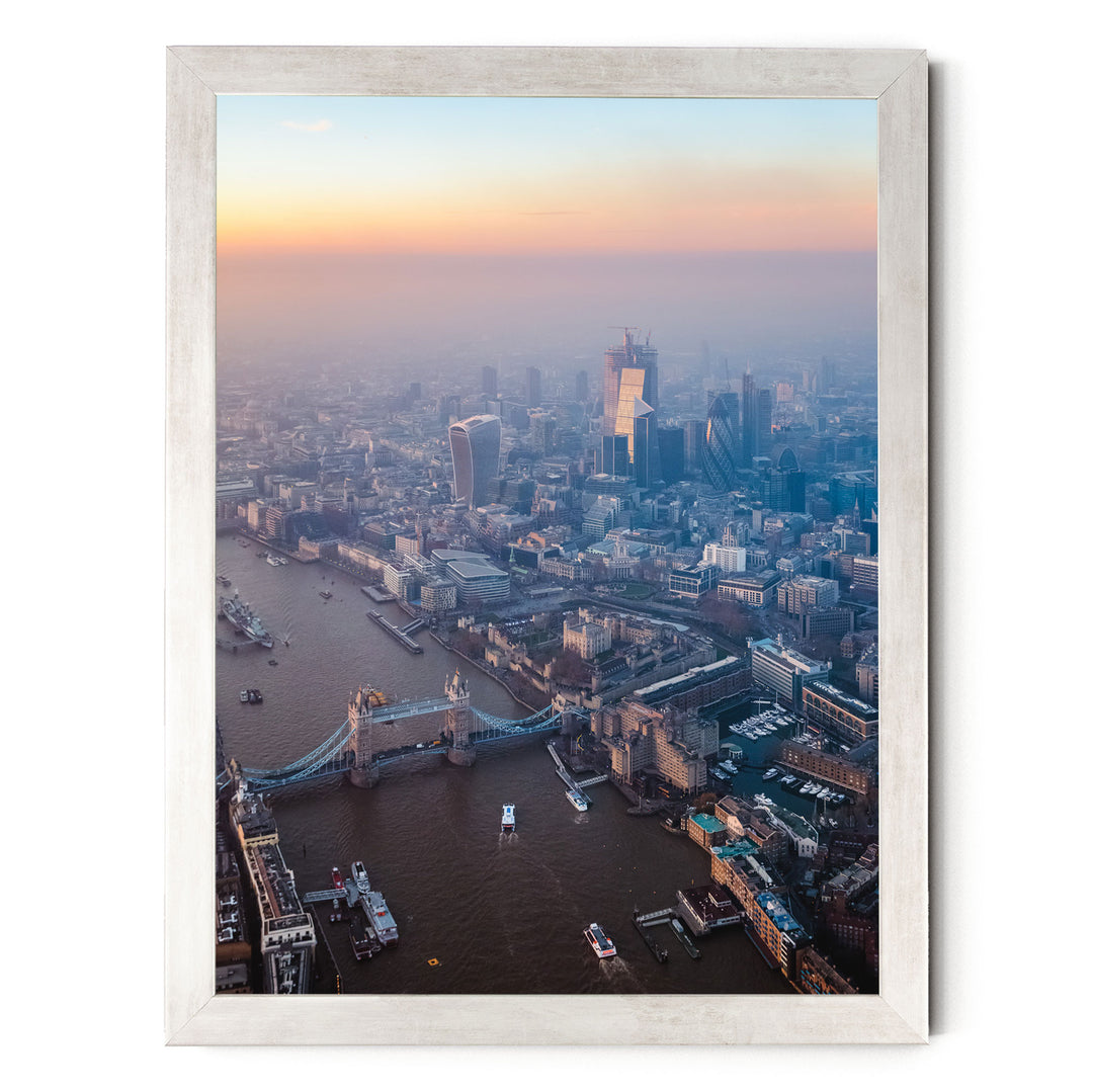 The image is an aerial view of a cityscape with a river running through it, featuring a bridge and several boats. The city is densely populated with tall buildings, and the sky is painted in warm hues of orange and pink, suggesting either sunrise or sunset.