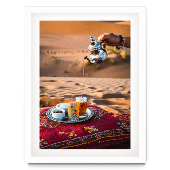 A hand pours tea from a silver teapot into a glass, with a tray of tea and snacks on a red and gold patterned cushion, set against a desert landscape.