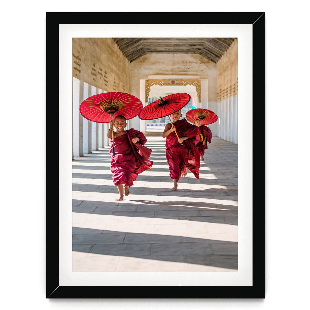 Two young monks in red robes are walking down a hallway, each holding a red umbrella.