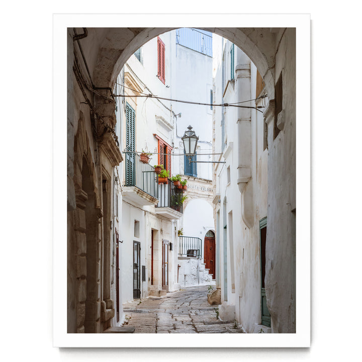 A narrow cobblestone street lined with white buildings, featuring arched doorways and balconies with flower boxes.
