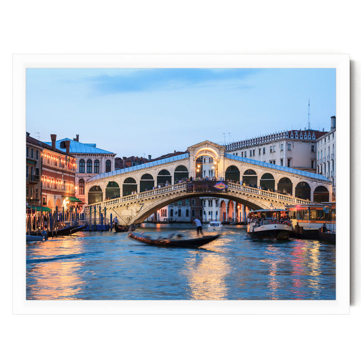a picturesque scene of the Rialto Bridge in Venice, Italy, with its distinctive white stone arch spanning the Grand Canal. The bridge is illuminated at dusk, creating a warm and inviting atmosphere. The canal is filled with boats, adding to the lively and bustling ambiance of the scene. The image captures the essence of Venice's iconic landmark, showcasing its architectural beauty and the vibrant life of the city.