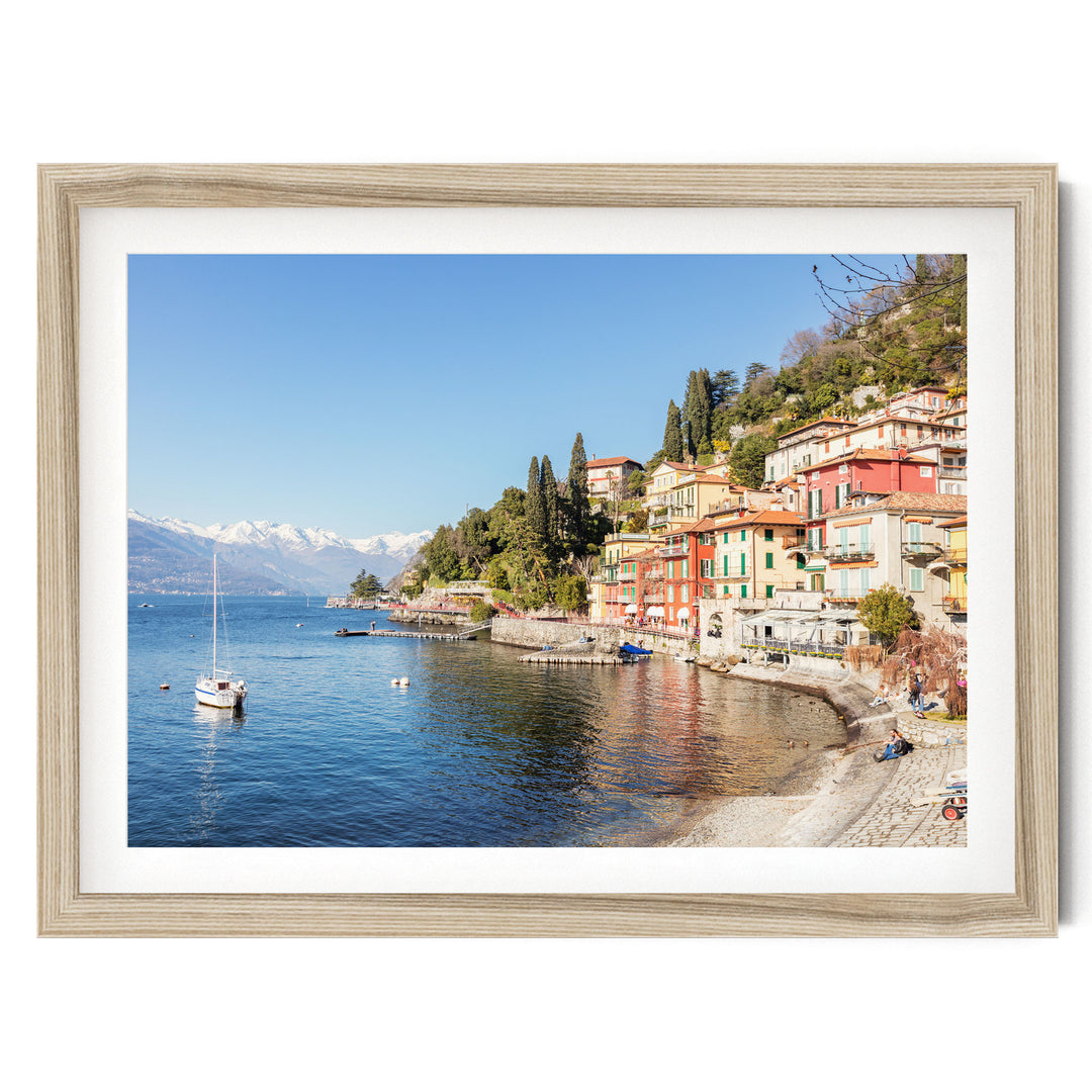 a picturesque coastal town with colorful buildings lining the shoreline, overlooking a serene lake. A sailboat is visible on the water, and the town is nestled among lush green hills.