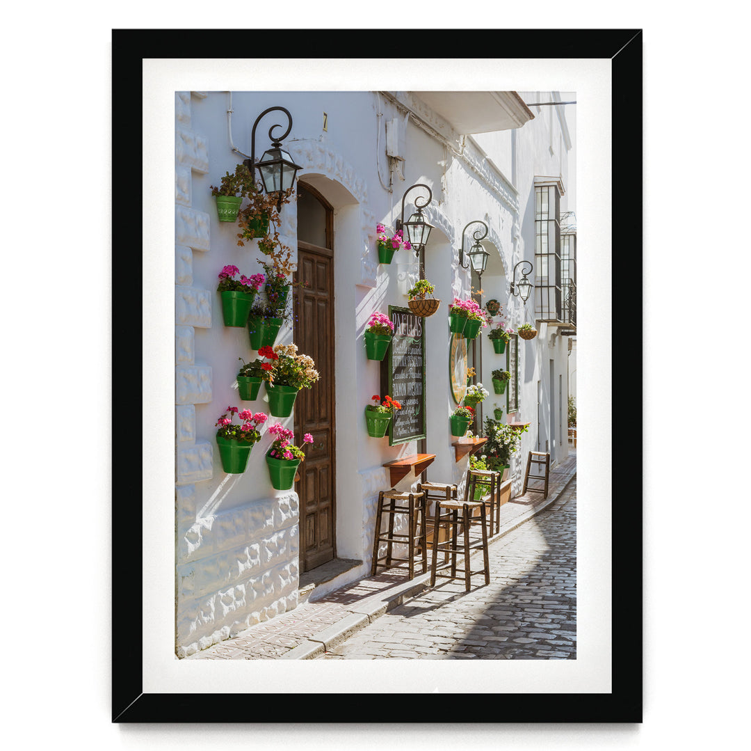 A charming street scene with a white building adorned with hanging flower pots and a wooden door. The building features a balcony with a wrought iron railing. The cobblestone street is lined with wooden stools and tables, creating a cozy outdoor seating area.