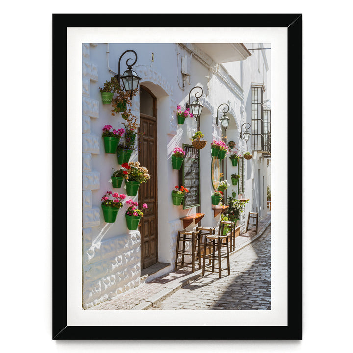 A charming street scene with a white building adorned with hanging flower pots and a wooden door. The building features a balcony with a wrought iron railing. The cobblestone street is lined with wooden stools and tables, creating a cozy outdoor seating area.