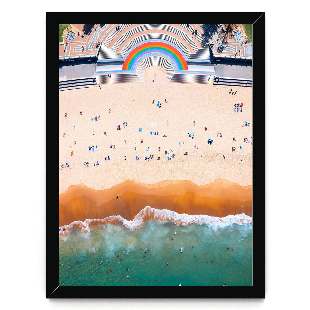 The image is an aerial view of a beach with a large rainbow arching over the top of it. The beach is crowded with people enjoying the sunny day, and the ocean waves can be seen crashing onto the shore.