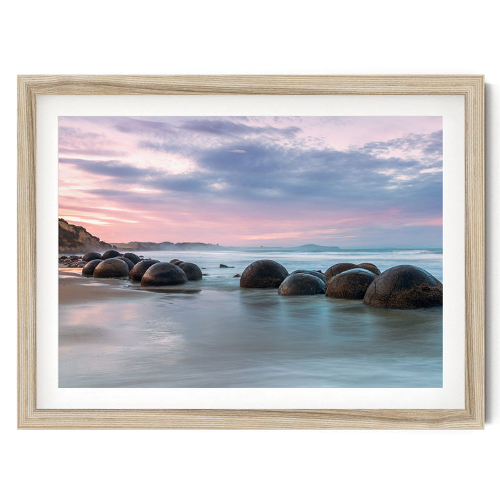 A serene beach scene with a collection of large, round boulders in the water, set against a picturesque sky with pink and purple hues.