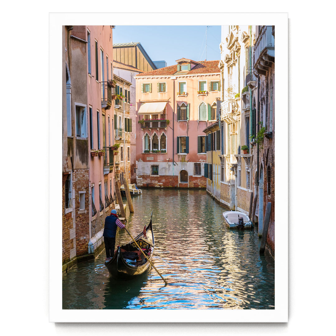 A gondolier in a traditional Venetian gondola navigates a picturesque canal in Venice, Italy, surrounded by colorful buildings and reflecting the clear blue sky.