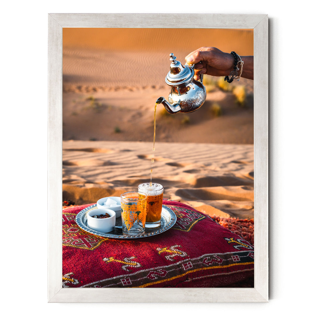 A person is pouring tea from a silver teapot into two glasses on a tray, set on a red and gold patterned cushion, with a desert landscape in the background.