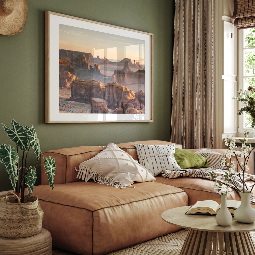 a living room with a large framed picture of a desert landscape hanging on the wall above a brown leather couch. The couch is adorned with throw pillows and a blanket, creating a cozy and inviting atmosphere. The room also includes a coffee table with a book and a vase of flowers, adding a touch of elegance and warmth to the space.