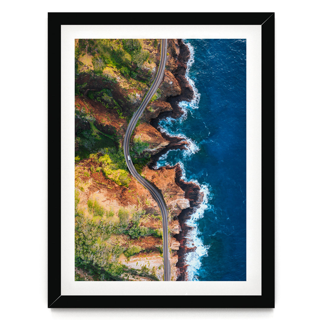 An aerial view of a winding coastal road cutting through a rugged, rocky coastline with crashing waves.